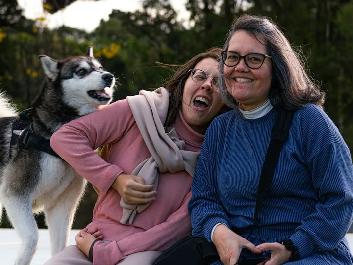 Two friends laughing on a bench outdoors with their husky leaning in